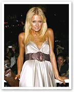 Blonde woman in a silver dress with a brown belt, standing in a crowded event.