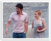 A man and woman walking closely on a cobblestone street, the woman holding a red clutch, in casual attire.