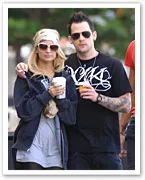 A couple in casual attire, wearing sunglasses, holding drinks, and standing close together outdoors.