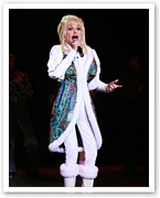 Singer performs on stage in vibrant attire with colorful vest, white outfit, and microphone in hand.