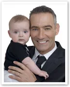 A man in a suit smiles while holding a baby, both looking towards the camera.