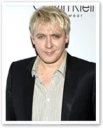 Man with blonde hair in a black suit and striped shirt, posing at a formal event.