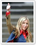 A woman with blonde hair raises her thumb in a gesture of approval, wearing a red shirt and dark jacket, standing outdoors.