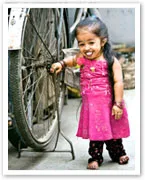 A smiling girl in a bright pink dress stands beside a bicycle wheel, reaching out to touch the spokes.