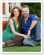 A couple sitting on grass, smiling, in casual attire with a garden in the background.