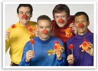 The Wiggles wearing colorful outfits and red clown noses, holding orange toy figures with red hair.