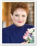 Portrait of a woman with short red hair, wearing a blue sweater, sitting beside colorful flowers.