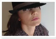 A person wearing a black hat and patterned shirt gazes upward with a neutral expression against a light background.