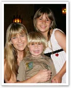 A woman and two children smile warmly for the camera, with one child wearing a khaki uniform and patches.