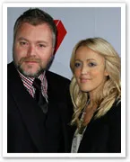 A man and woman posing together at an event, both dressed in formal attire with neutral expressions.
