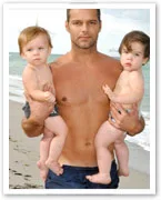Man holding two toddlers on a beach, shirtless, with the ocean in the background.