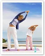 Mother and child doing side stretches on yoga mats outdoors, with ocean in background.