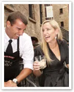 A woman in a black dress and a man in a white shirt share a laugh outdoors, she holds a glass, brick building in the background.