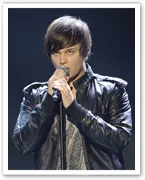 Young man with brown hair singing into a microphone, wearing a black leather jacket on stage.