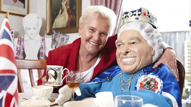 Person with statue resembling a monarch at a table with tea cups and a Union Jack flag, smiling and holding a themed cup.
