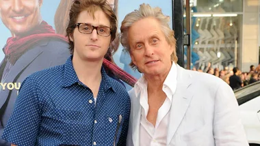 Two men posing at an outdoor event, one in a blue shirt with glasses, the other in a white suit, with a crowd in the background.