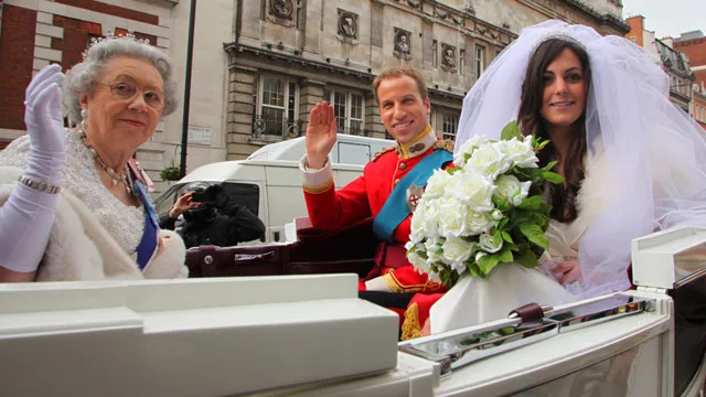 Fake royals in a carriage: an elderly woman, man in soldier uniform, and woman in wedding dress waving to crowd.