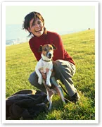A person kneeling on grass, smiling, and holding a small dog.