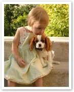 A young child in a light dress sits on steps, gently holding a small brown and white puppy, with greenery in the background.