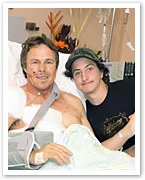 In a hospital bed, a smiling man with arm in sling poses with a young man standing beside him.