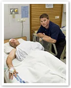 Two men in a hospital room; one is lying in bed, bandaged with an IV, the other is standing beside him, looking concerned.