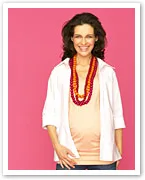 Pregnant woman smiles, wearing colorful necklaces over a light shirt and white overlay, with a pink background.