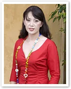 Jacqueline Pascarl wearing a red dress and colorful necklace, standing against a beige wall with greenery.