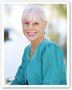 Senior woman with white hair smiling while sitting outdoors in a turquoise blouse.