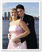Couple in formal attire smiling by a waterfront with city skyline in the background.