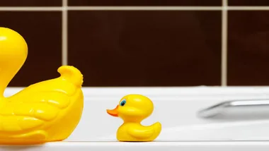 Yellow rubber ducks sit on the edge of a white bathtub against a tiled brown wall.