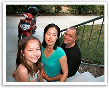 Family of three smiling on outdoor steps with a red scooter in the background.