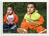 Two toddlers in colorful outfits sitting on grass, one in orange and one in red, looking towards the camera.