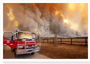 A fire truck is parked near a fence with a large bushfire burning in the background, producing thick smoke.