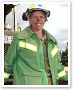 Female firefighter in green protective gear and helmet, smiling, with a firetruck in the background.