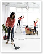 Four women in red shirts doing household chores: vacuuming, fixing windows, folding laundry, and dusting in a bright room.