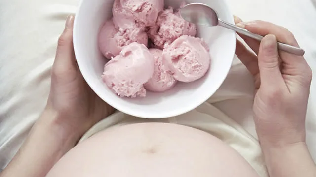 Pregnant person holding a bowl of pink ice cream, spoon in hand.