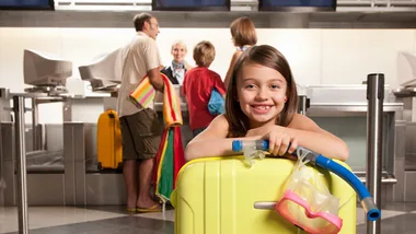 Child smiling at airport check-in with suitcase and snorkel gear, family in background talking to airline staff.