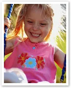 A young girl in a pink dress with floral design is smiling while swinging outside on a sunny day.