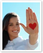 A smiling person holds up a hand with a red heart painted on the palm against a clear blue sky background.