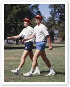 Two people power walking in a park, wearing white shirts, blue shorts, red caps, and white sneakers.