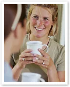 A smiling woman holding a white mug, sitting across from another person, engaging in conversation.