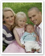 A family of four sits on grass: a woman, a man, a young girl in pink holding a sleeping baby, all smiling.