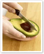 A person slicing an avocado's seed with a knife on a wooden cutting board.