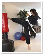 Female martial artist in black gi and white belt practicing a high kick on a red punching bag.