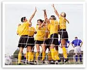 Soccer team in yellow jerseys huddling and giving high-fives on the field, netting visible in the foreground.