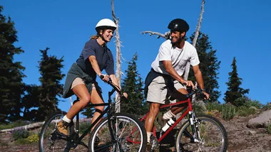 Two people mountain biking on a trail, smiling in sunny weather, surrounded by trees and a clear blue sky.