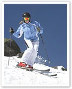 Skier in blue jacket and white pants skiing downhill on a sunny day, with snow and rocks in the background.