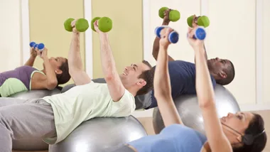 A group of people exercising on stability balls, lifting dumbbells overhead in a fitness class setting.