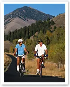 Two people biking on a paved trail with mountains and trees in the background on a sunny day.