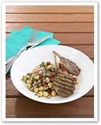 Grilled lamb chops with a chickpea salad on a white plate, set on a wooden table with a teal napkin and cutlery.